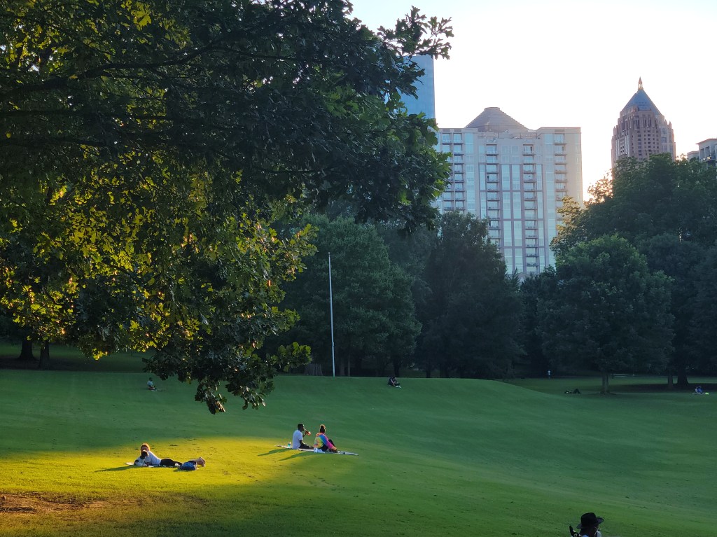 People lounging on a grassy field in a park with a skyline in the background during sunset.