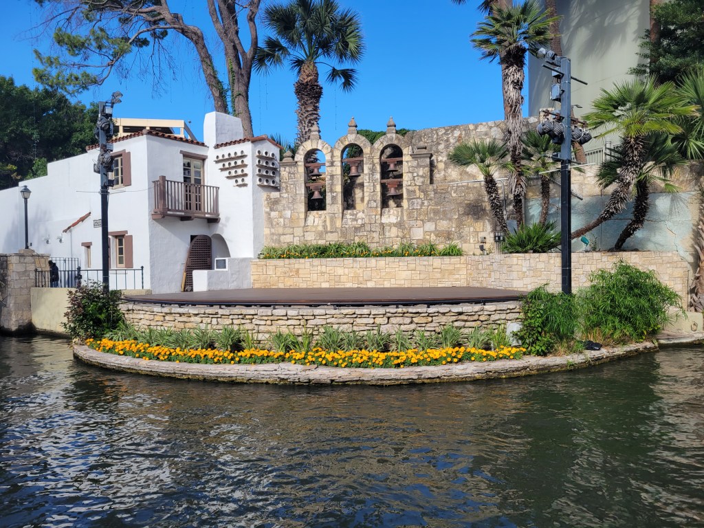 A section of the San Antonio River Walk featuring a scenic riverside stage surrounded by greenery and vibrant orange flowers, with a backdrop of a stone wall and palm trees under a clear blue sky.