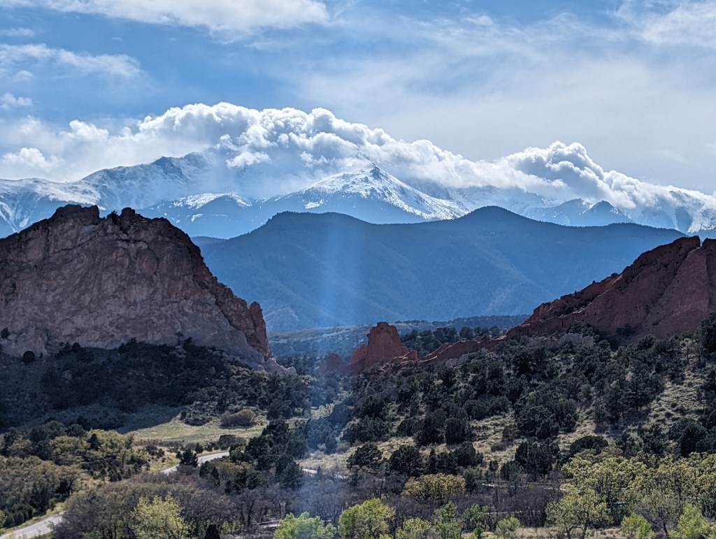 A scenic view of rocky terrain in the foreground with lush greenery, leading to mountains covered in snow and clouds in the background, under a bright blue sky.